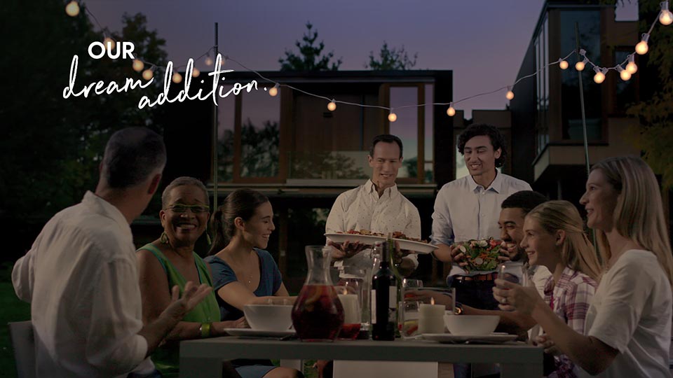 A couple serves guests in their backyard. Everyone is smiling and enjoying themselves on this summer's night. The backyard is beautiful with fairy lights throughout. Text says "our dream addition".
