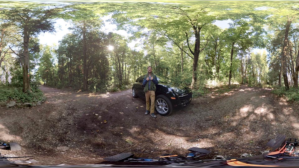 A young man stands in front of a Porsche car in the middle of a forest.