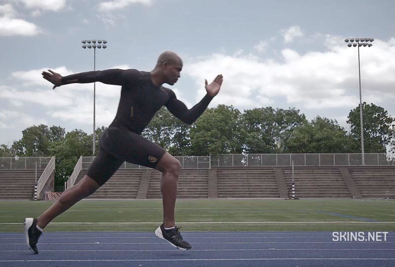 A sprinter in SKINS athletic wear is caught in a mid-start stride in a large outdoor track stadium.