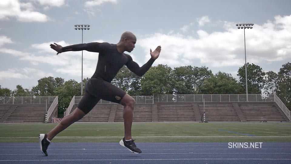 A sprinter in SKINS athletic wear is caught in a mid-start stride in a large outdoor track stadium.