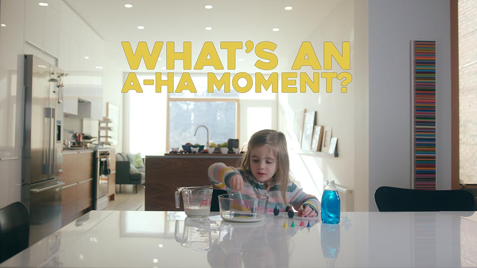A little girl places a Q-tip into a bowl of milk and food colouring - conducting a small science experiment. She is in a big open kitchen. Above her is the text "What's an a-ha moment?"