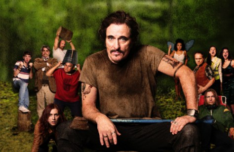 Canadian icon Kim Coates poses for a theatre poster on a wooden stump with an axe in his hand. Behind him are the 13 additional members of the cast, in full party mode. The setting around them is a forest.