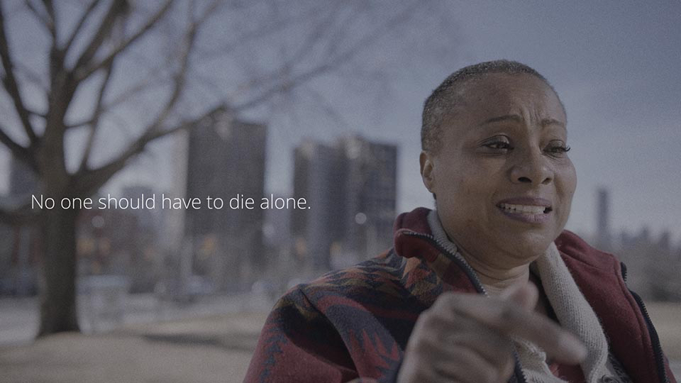 A woman speaks tearfully directly to camera, with the text-on-screen saying "No One should have to die alone."